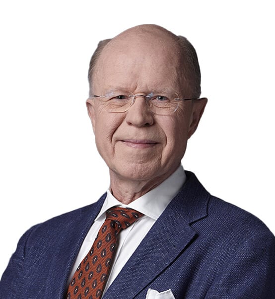 An older man with glasses wearing a blue suit, white shirt, and patterned brown tie, smiling slightly. The background is plain white.